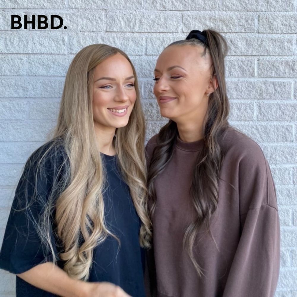 Two smiling women wearing curly BHBD wigs: one with a blonde wig and the other with a brown wig.