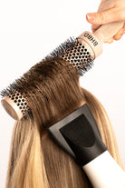 Close-up of a BHBD round hairbrush lifting a section of blonde hair while a hairdryer nozzle applies heat, demonstrating a professional blow-drying technique for smooth and voluminous results.